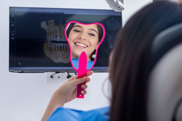 Optimistic teen patient smiling and looking at teeth in mirror against monitor with denture model during check up appointment in clinic.